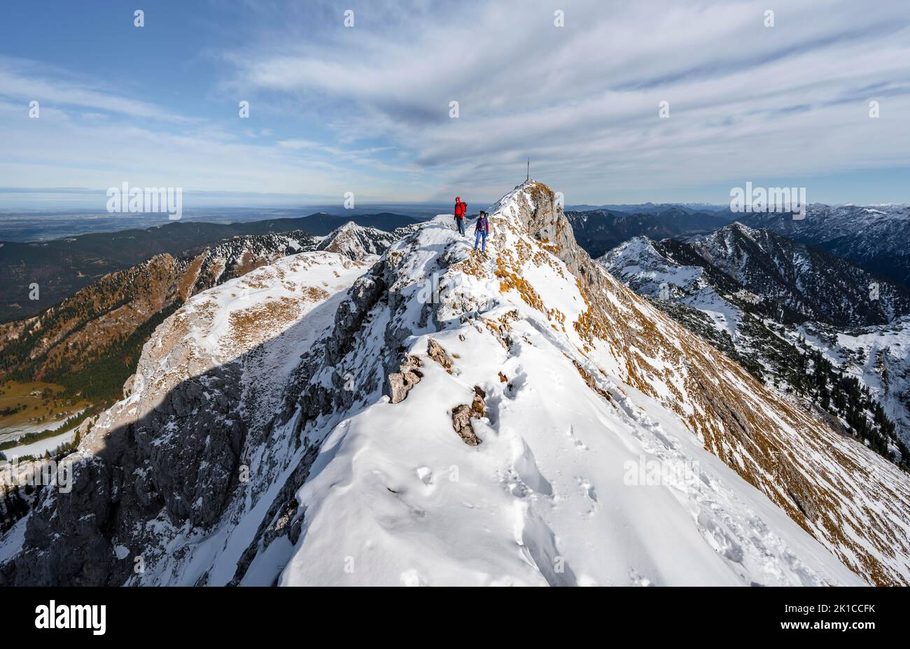 Two climbers on a narrow rocky snowy ridge, summit of the Ammergauer Hochplatte with summit cross, view of mountain panorama, hiking in autumn Stock Photo