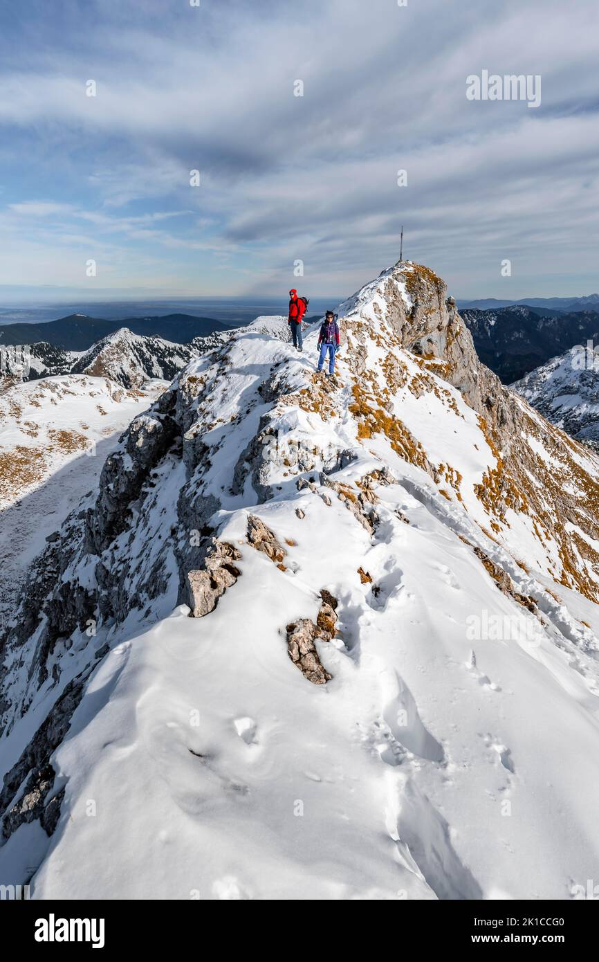 Two mountaineers on a narrow rocky snowy ridge, in the back summit of the Ammergauer Hochplatte with summit cross, view of mountain panorama, hiking Stock Photo