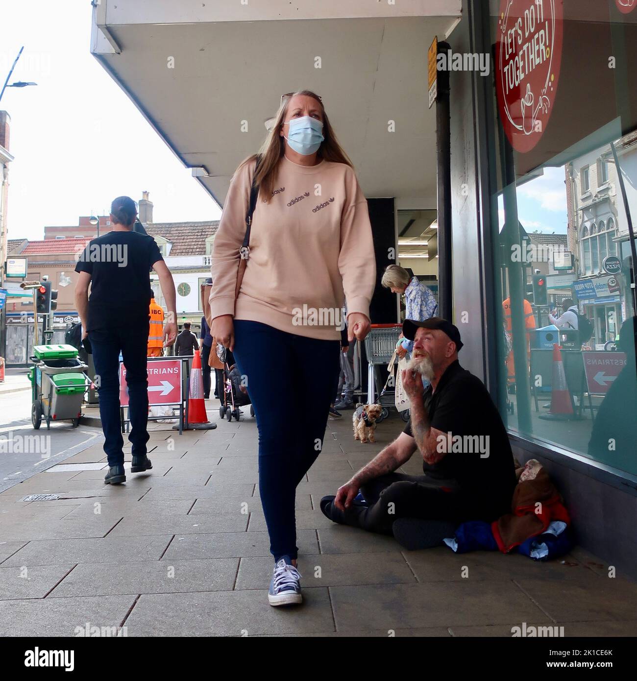 Ipswich, Suffolk, UK - 17 September 2022 :A woman wearing a mask walks past a beggar seated on the ground outside a supermarket. The beggar is asking for spare change. The woman ignores him. Stock Photo