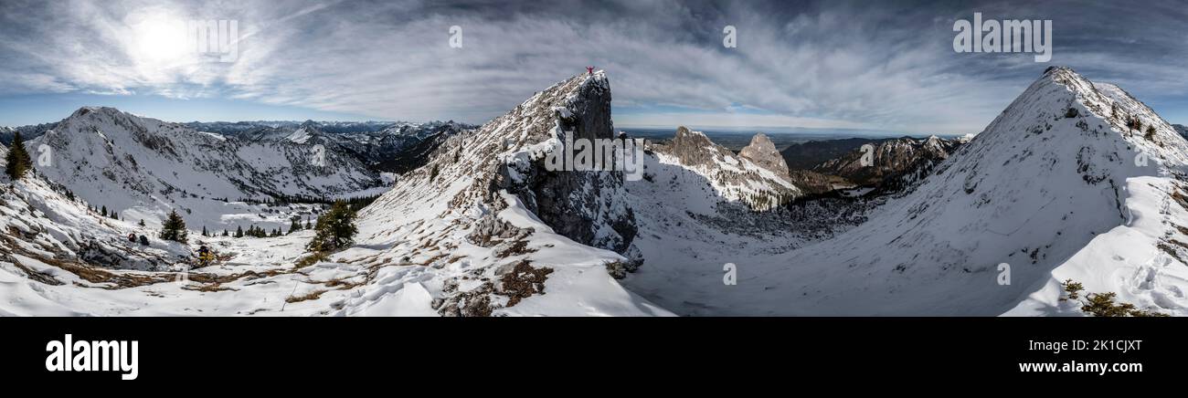 Mountaineer stretches his arms in the air, standing on a snowy rocky ridge, window on the hiking trail to Ammergauer Hochplatte, in autumn, panorama Stock Photo