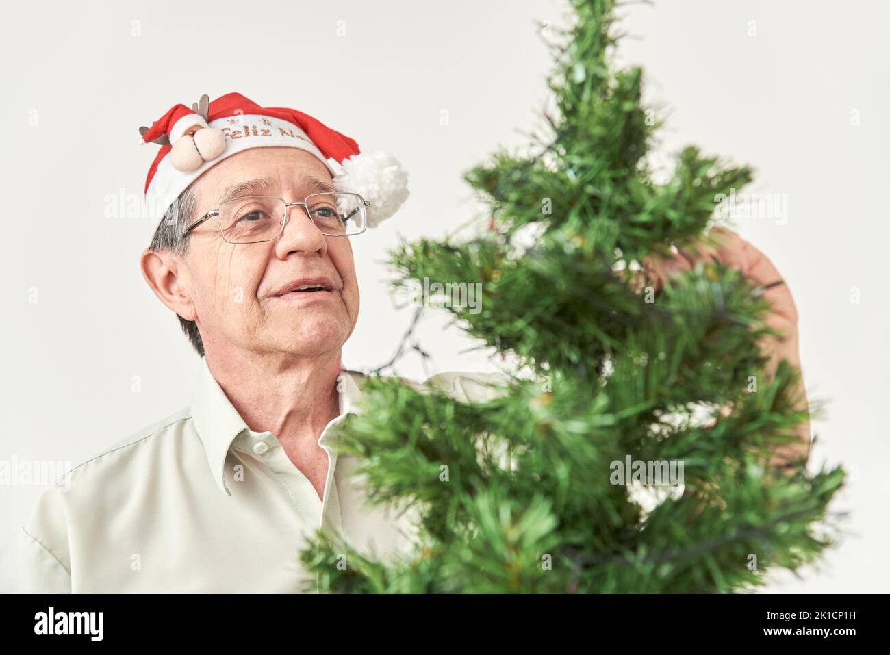 Happy senior hispanic man smiling while decorating a Christmas tree at home wearing a red Santa Claus hat with the text Merry Christmas. The happiness Stock Photo