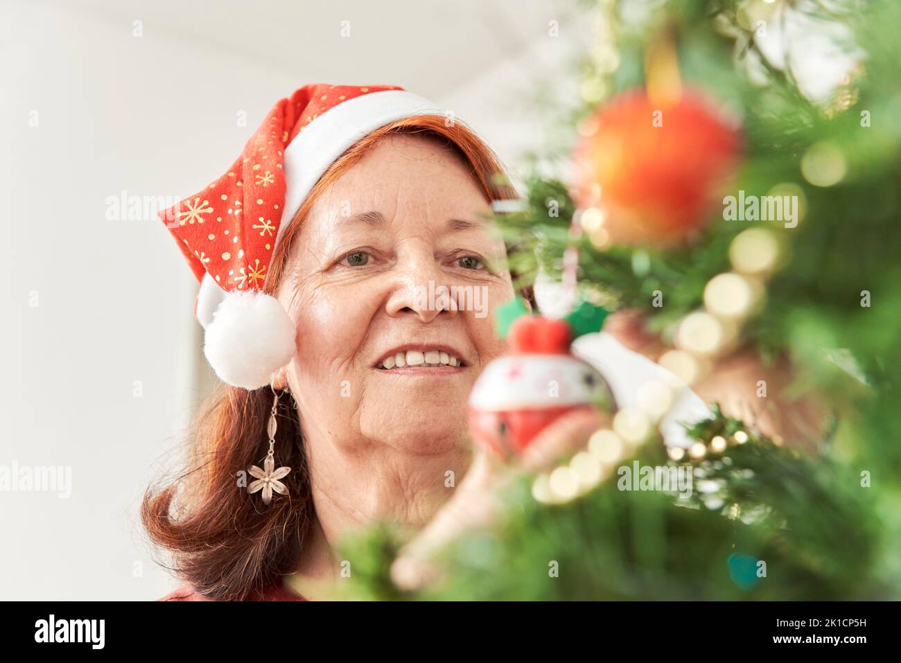 Happy senior hispanic woman smiling while decorating a Christmas tree at home wearing a red Santa Claus beanie. Stock Photo