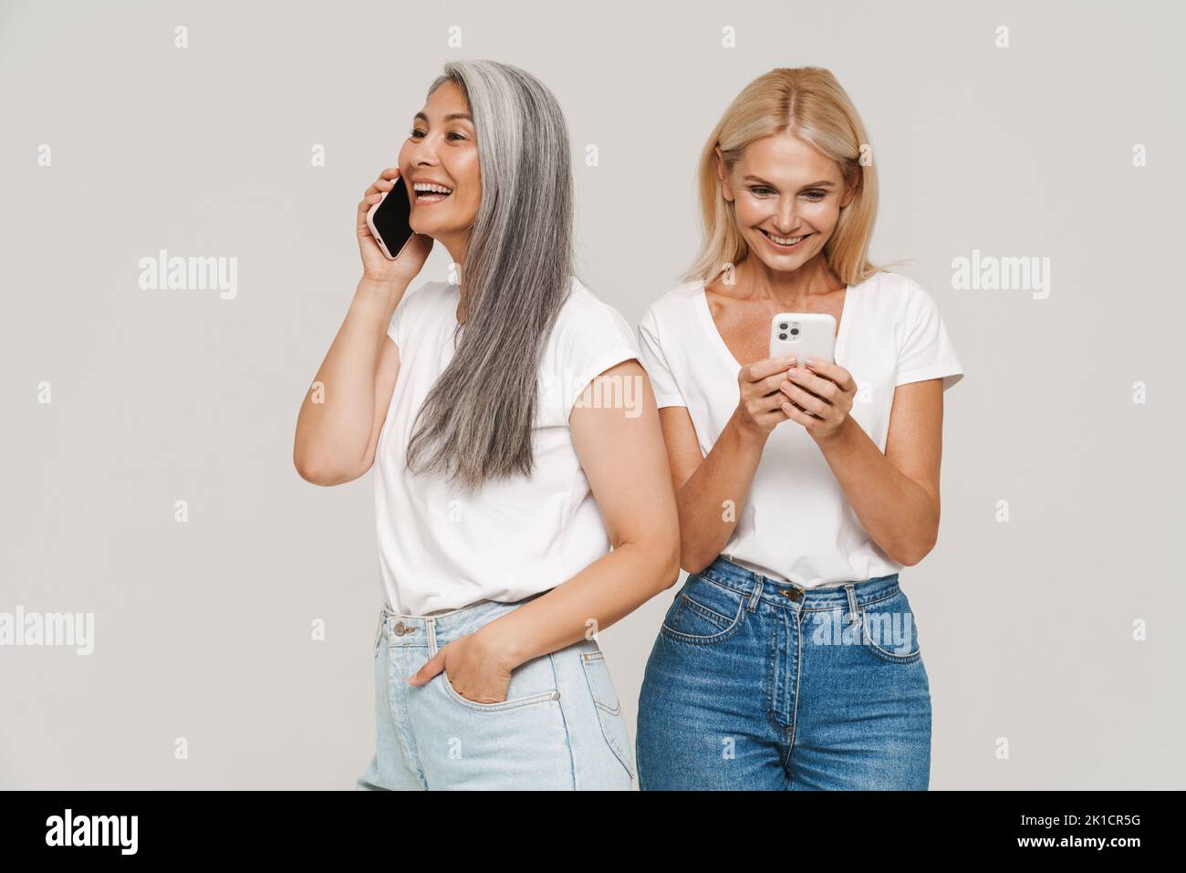 Mature multiracial women wearing t-shirts smiling and using cellphones together isolated over white background Stock Photo