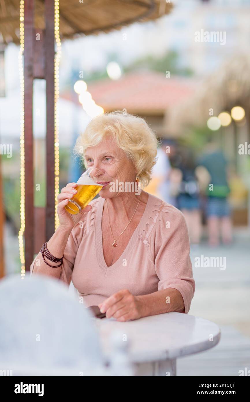 Mature woman sitting at a table in a summer cafe and drinking beer Stock Photo