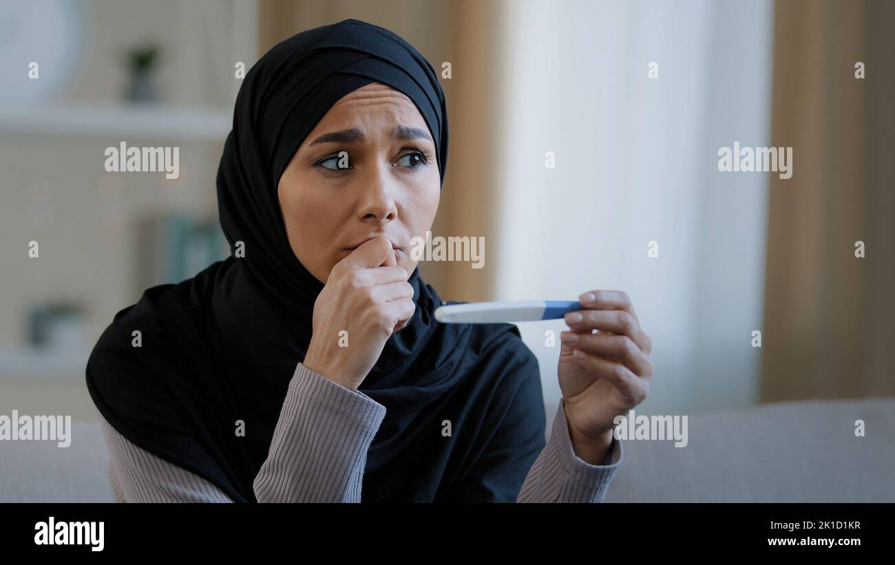 Shocked unhappy islamic young woman arabian muslim girl in hijab female adult pregnant feeling fear looking at positive pregnancy test result sit at Stock Photo