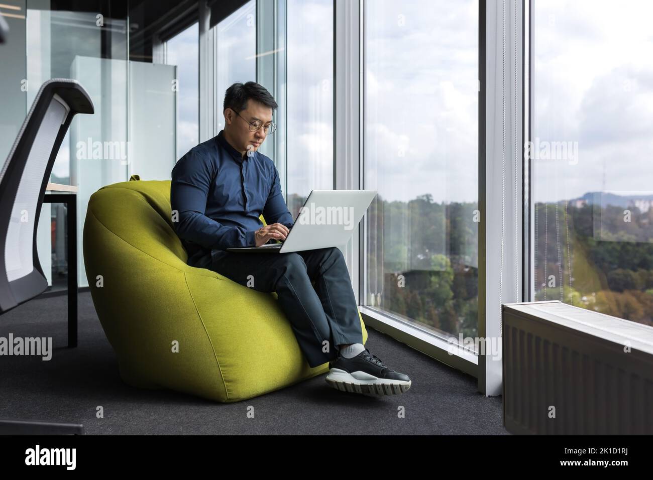 Asian young office worker sitting on soft ottoman chair, programmer using laptop for writing code and programming testing new program, man sitting near window inside office. Stock Photo