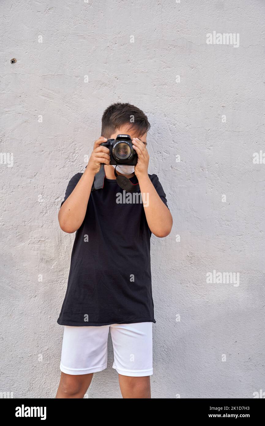 A cute little kid with green eyes wearing a mask is holding a camera and taking pictures . Happy boy dreams of becoming photographer. Stock Photo