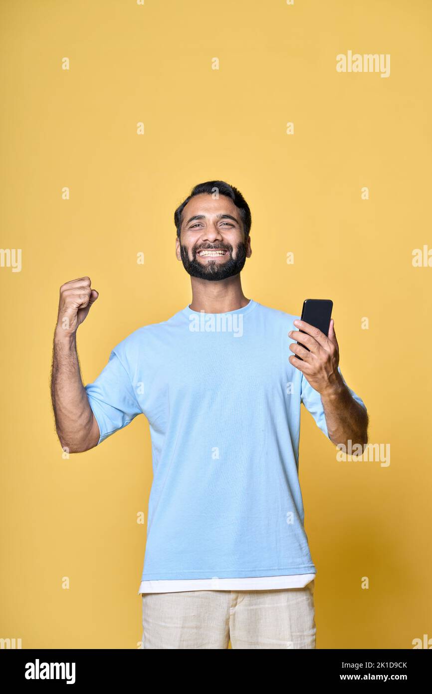 Excited indian man raising fist using phone isolated on yellow, vertical. Stock Photo