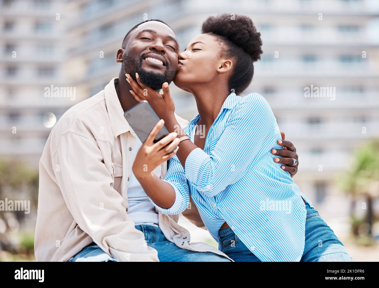 Love, kiss and couple take phone selfie on romantic date together, happy and smile while relax outdoors on summer holiday. Black woman and man on a Stock Photo