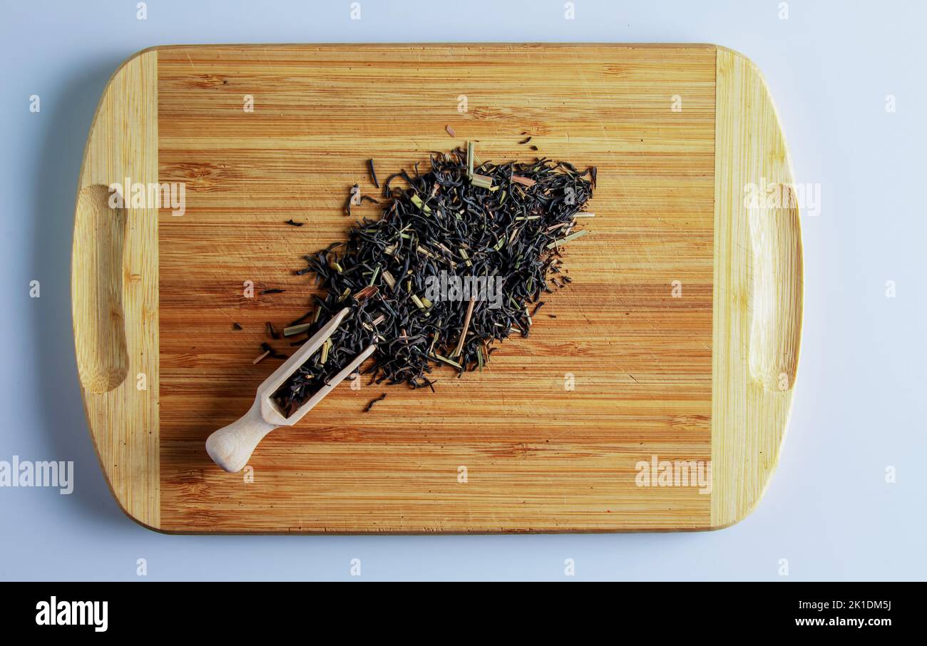 The tea is ready to be brewed. Spatula for brewing tea. Background of fragrant black tea leaves with lemon grass. Tea on a wooden board. Stock Photo