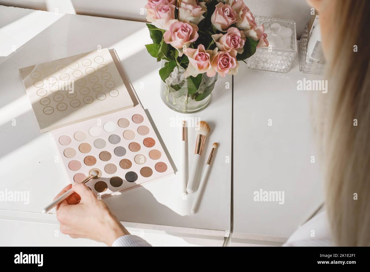 Hand taking cosmetics for make-up, roses flowers on table. Decorative eyes shadows with foundation on dressing table in beauty salon. Artist choosing Stock Photo