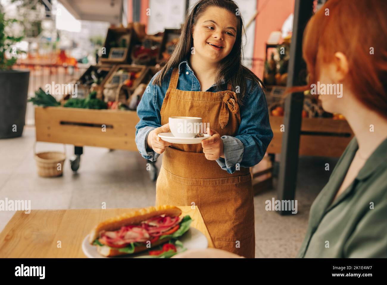 Friendly waitress with Down syndrome serving a customer a sandwich and coffee in a trendy cafe. Professional woman with an intellectual disability wor Stock Photo