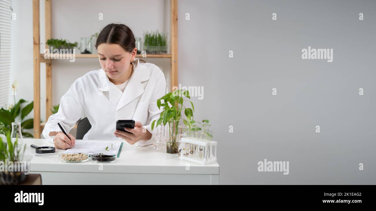 A young attractive woman scientist with protective gloves using a smartphone in the scientific bio laboratory, writing results on a clipboard. Growing Stock Photo