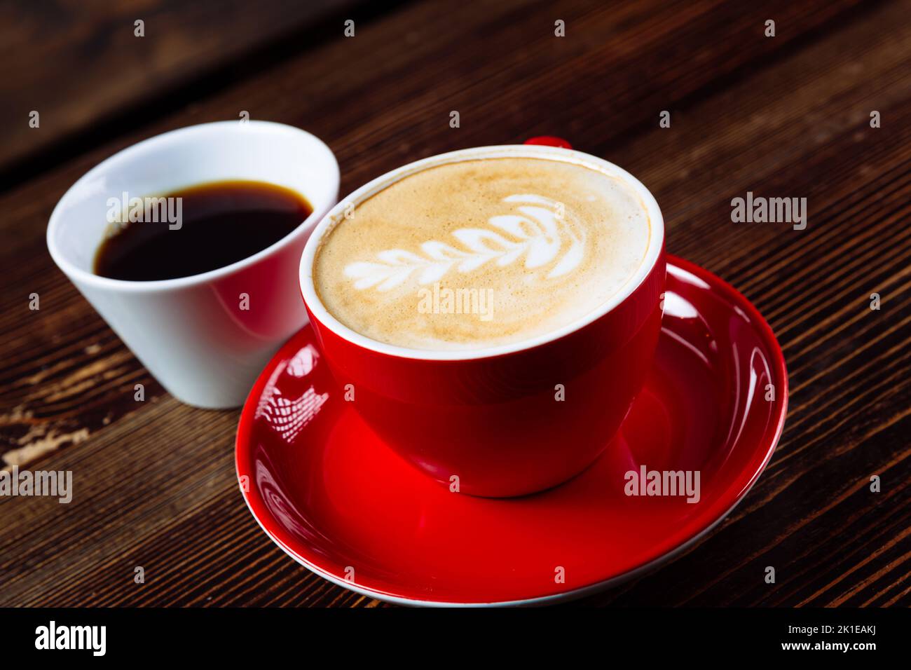 Aromatic coffee in a red cup with milk foam and latte art and freshly brewed coffee in a white cup on a light wooden table. Close up Stock Photo