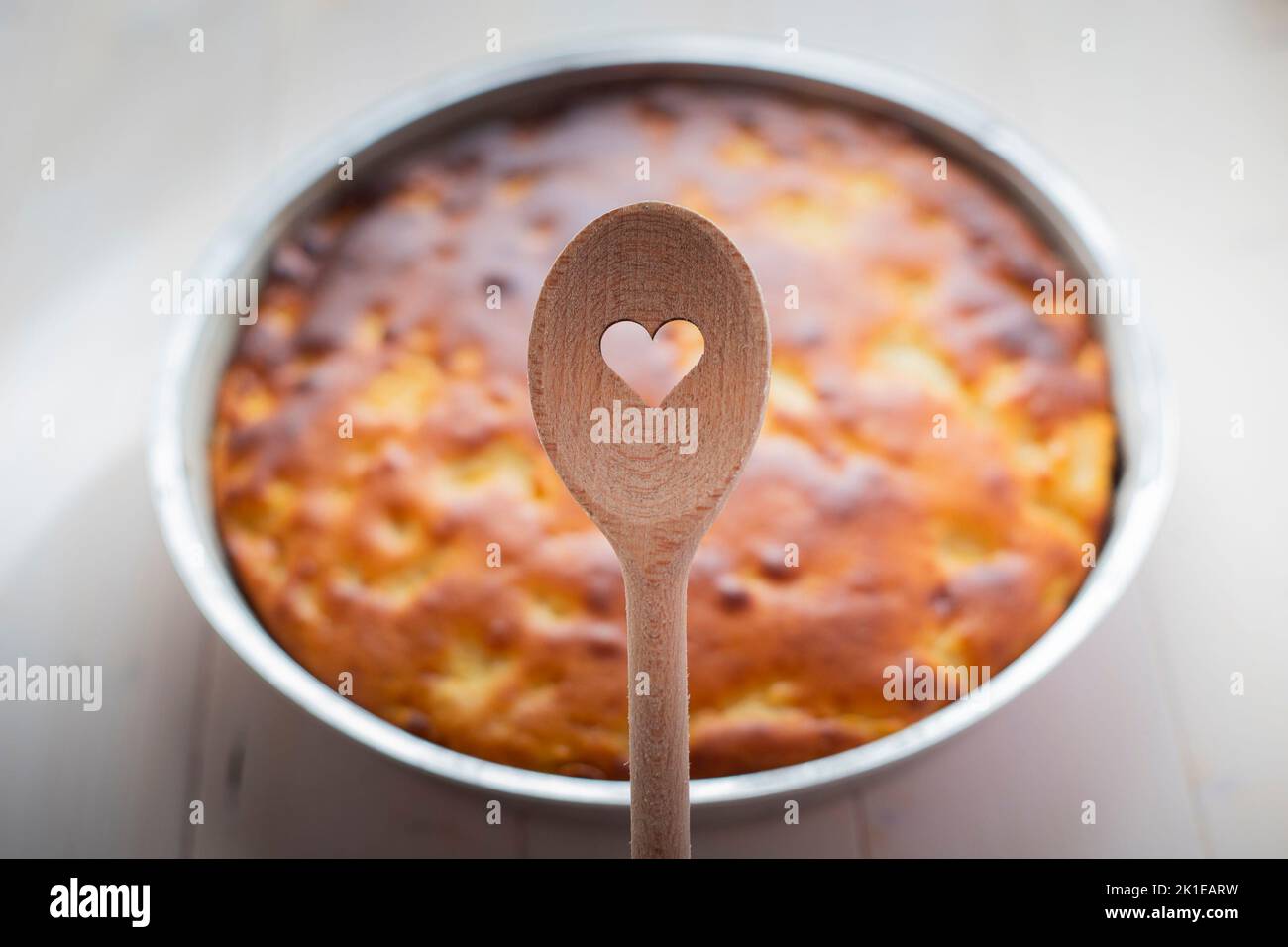 Wooden spoon with heart shaped hole, and pie in the background on white table. Baked with love concept. Stock Photo
