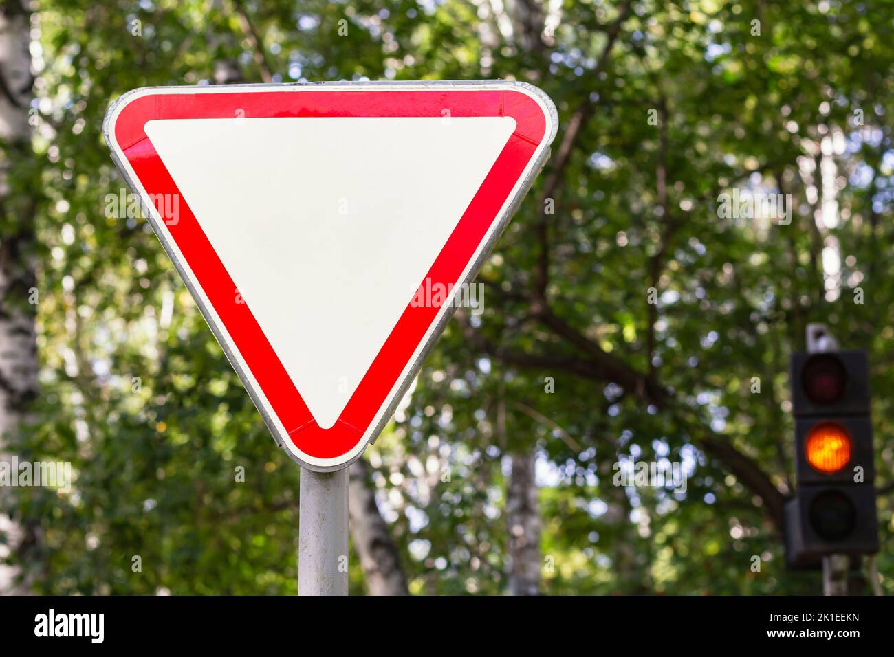 Russian road sign give priority to traffic on main road ahead. Give way sign with trees and traffic light in blur in background.Danger warning and sto Stock Photo