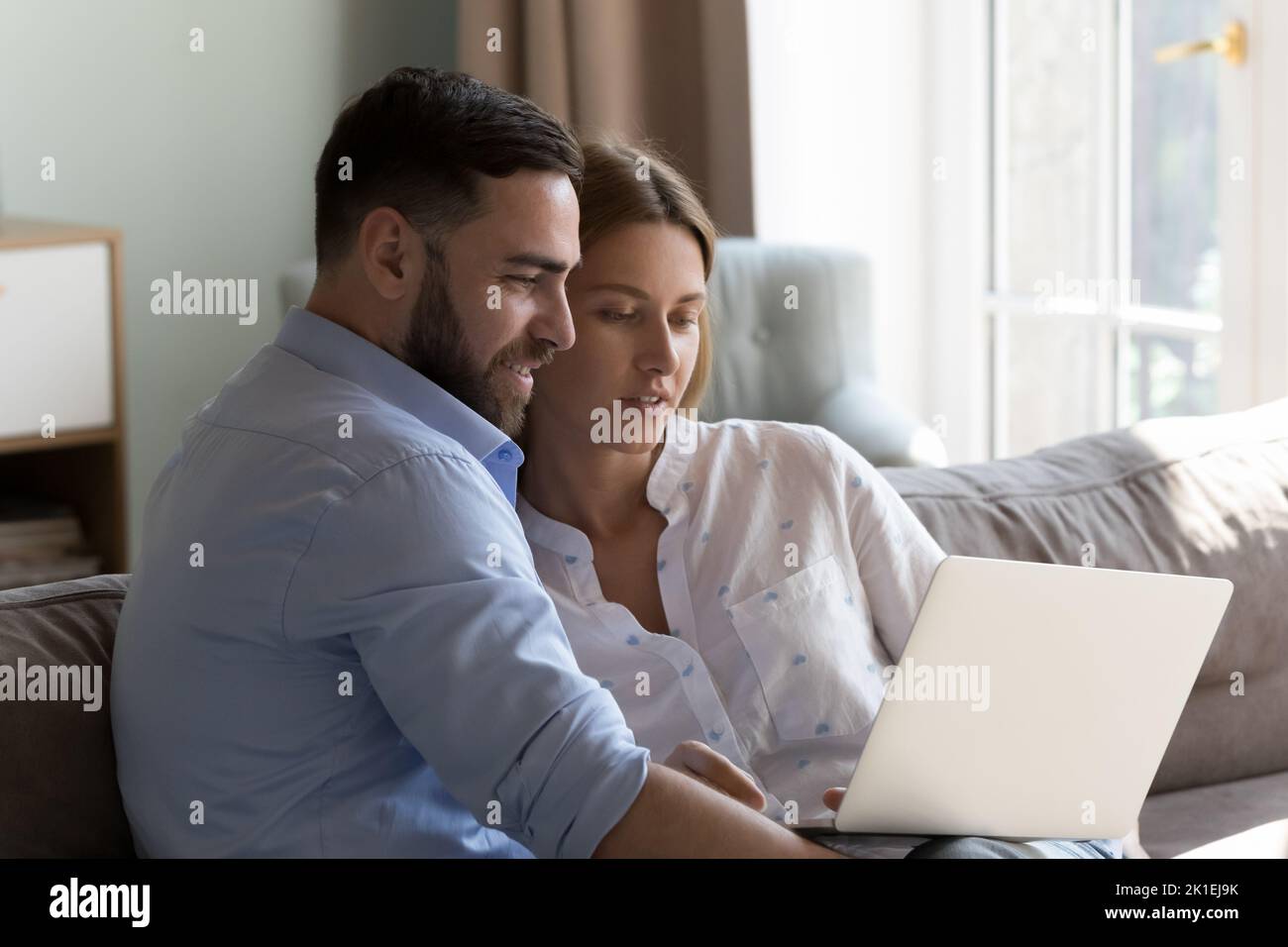 Happy millennial couple resting on sofa at home, holding laptop Stock Photo