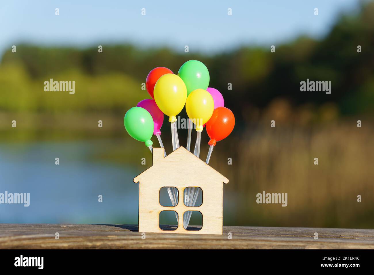 Miniature flat house shape with a bunch of small colorful balloons against a beautiful lake landscape background. Creative moving in party concept. Stock Photo