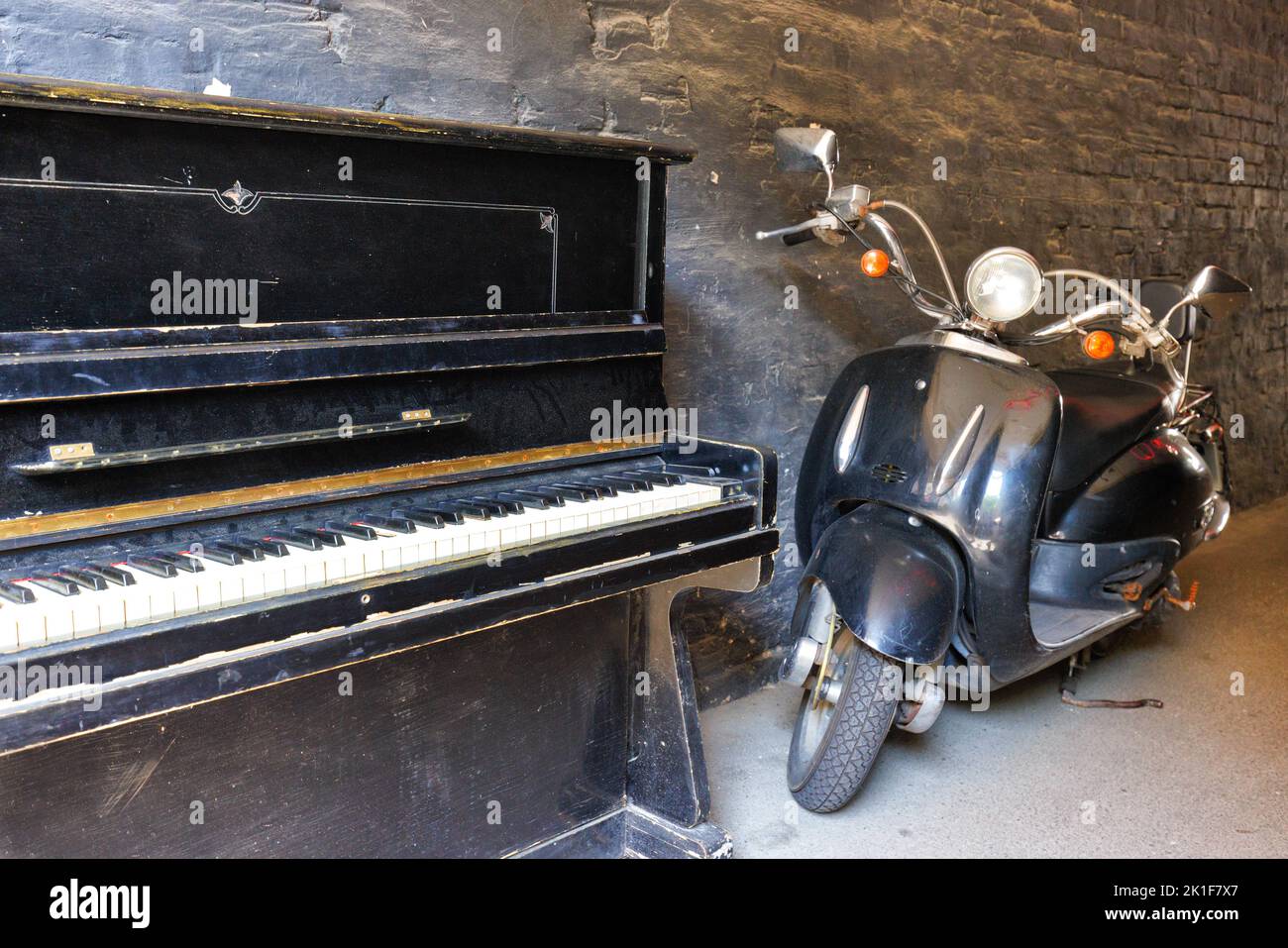 A retro piano near a vintage motorcycle against the background of an old brick wall painted with black paint. Stock Photo
