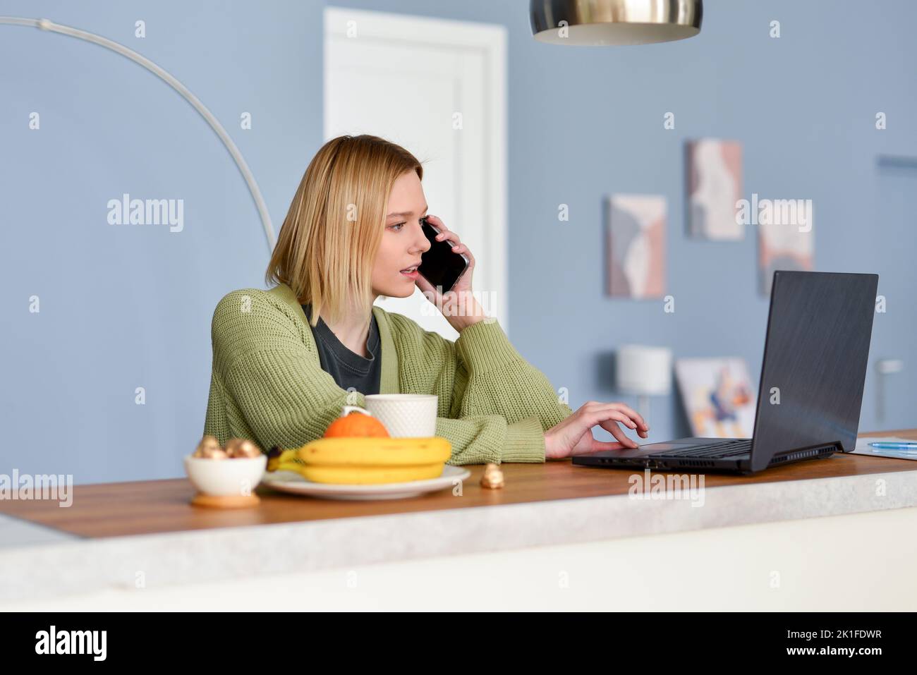 Young woman talking on the phone in the kitchen and using her laptop. Stock Photo