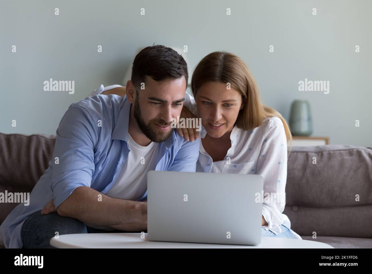 Focused positive millennial married couple sitting on couch at laptop Stock Photo