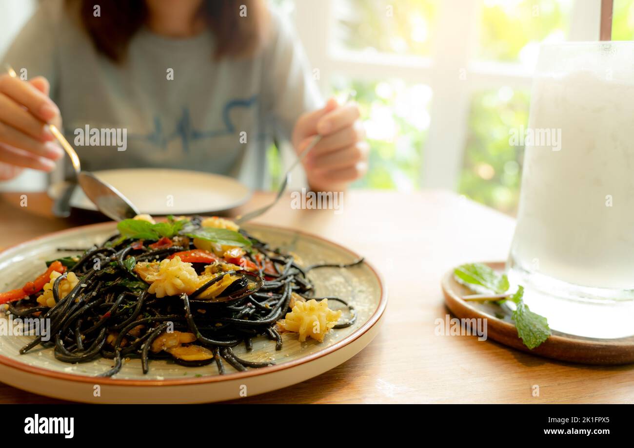 Black cuttlefish ink spaghetti with shrimp on plate. Black pasta with squid ink on a restaurant table and blur woman eating with fork and spoon. Stock Photo