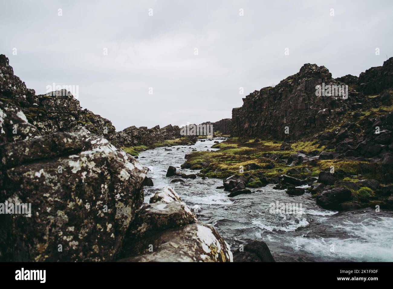 A mountain stream of dark cold water near the waterfall of Oxararfoss in Thingvellir National Park, Iceland. Green grass amidst the rocks beneath heav Stock Photo
