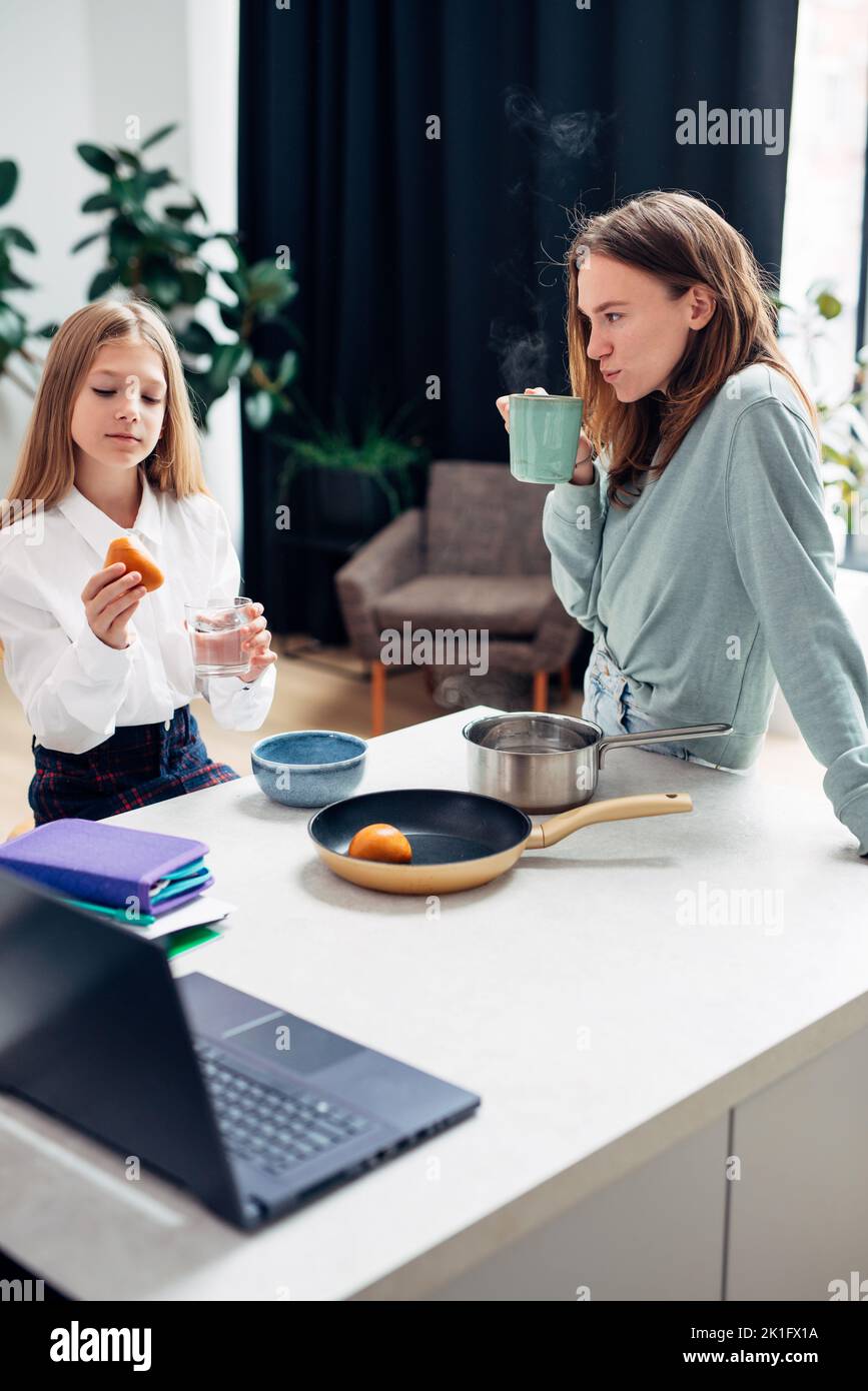 Girl is eating a bun and her mother is drinking coffee. Breakfast before school and work. Stock Photo