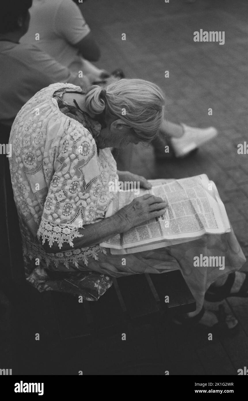 A vertical shot of an elderly woman reading a newspaper on a bench in grayscale Stock Photo