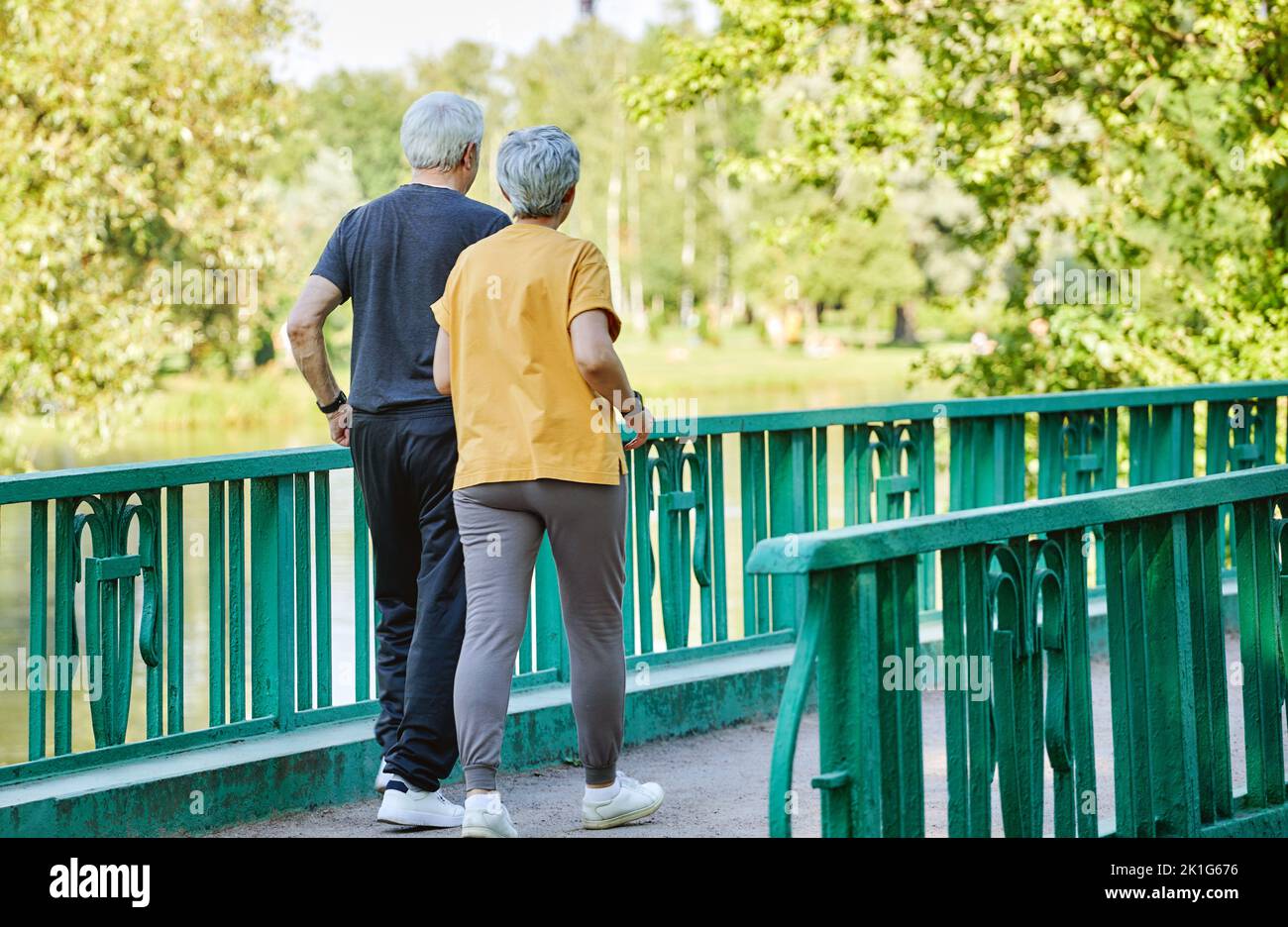 Rearview middle-aged wife and husband wear activewear strolling along bridge in summer park, enjoy morning sportive walk together outside. Healthy lif Stock Photo