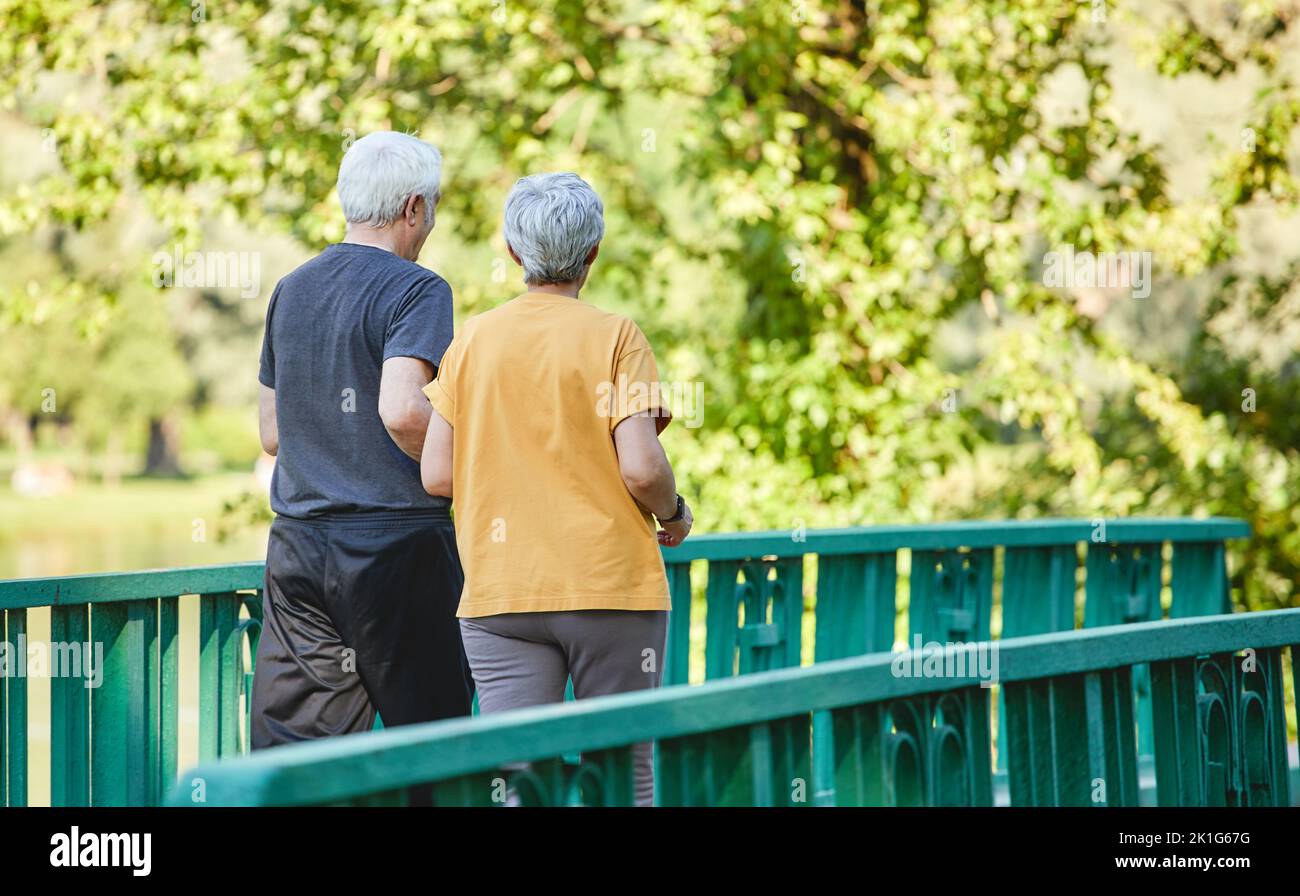 Rearview middle-aged wife and husband wear activewear strolling along bridge in summer park, enjoy morning sportive walk together outside. Healthy lif Stock Photo