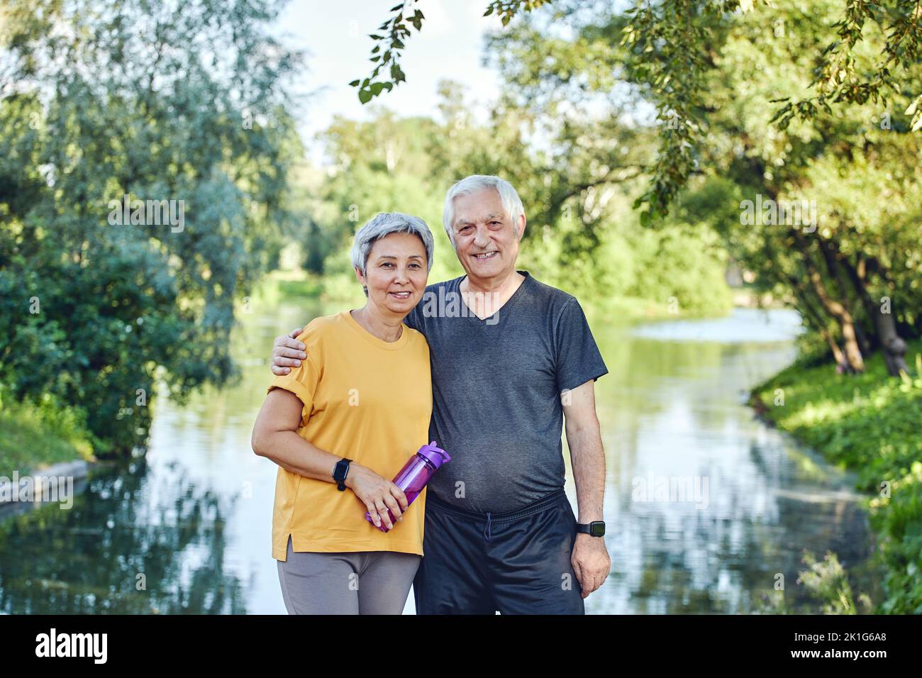 Attractive older couple in sportswear hugging pose look at camera standing in summer park near lake after morning stroll, sport active lifestyle holds Stock Photo