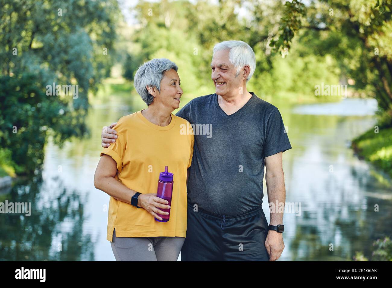 Attractive older couple in sportswear hugging pose look at each other standing in summer park near lake after morning stroll, sport active lifestyle h Stock Photo