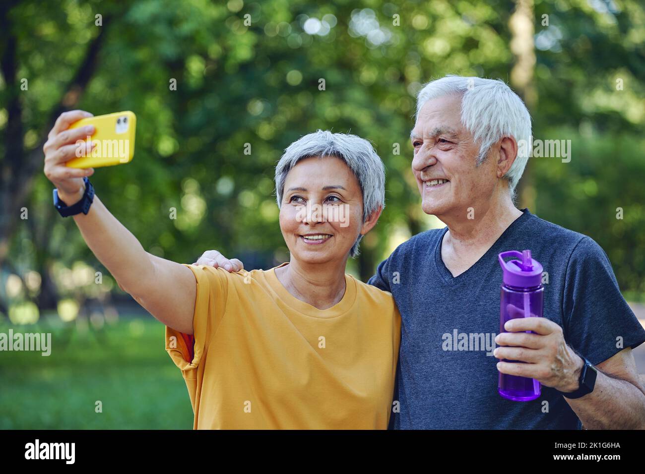 Old sportive couple staring at smart phone cam take selfie picture enjoy modern wireless tech usage while stroll in sportswear in summer park. Carefre Stock Photo