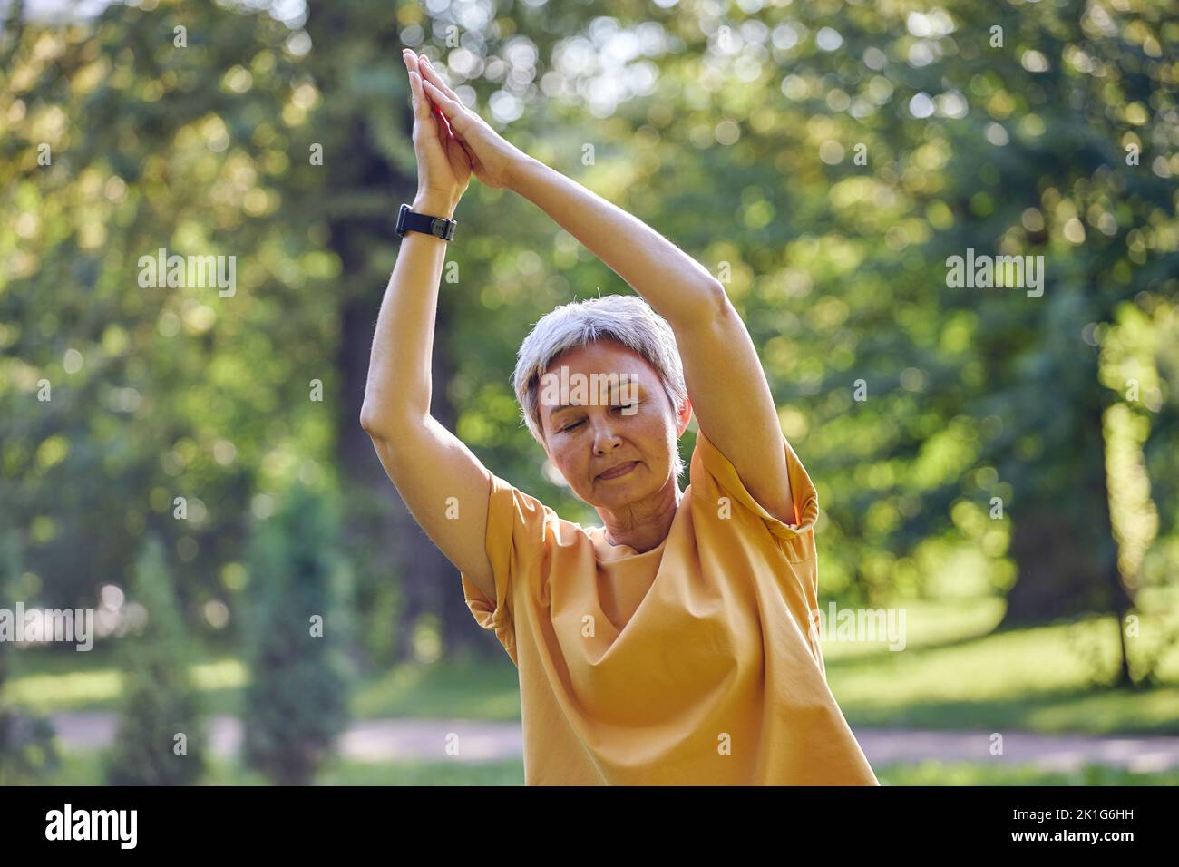 Older active Asian woman in sportswear close her eyes meditating in summer park with folded raised palms makes stretching exercises, do yoga work out Stock Photo