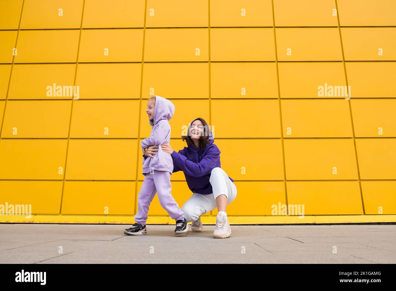 Woman in purple hoody have fun with cute girl 4-5 years old. Mommy and little daughter on yellow mall wall background. Childhood concept of love for Stock Photo