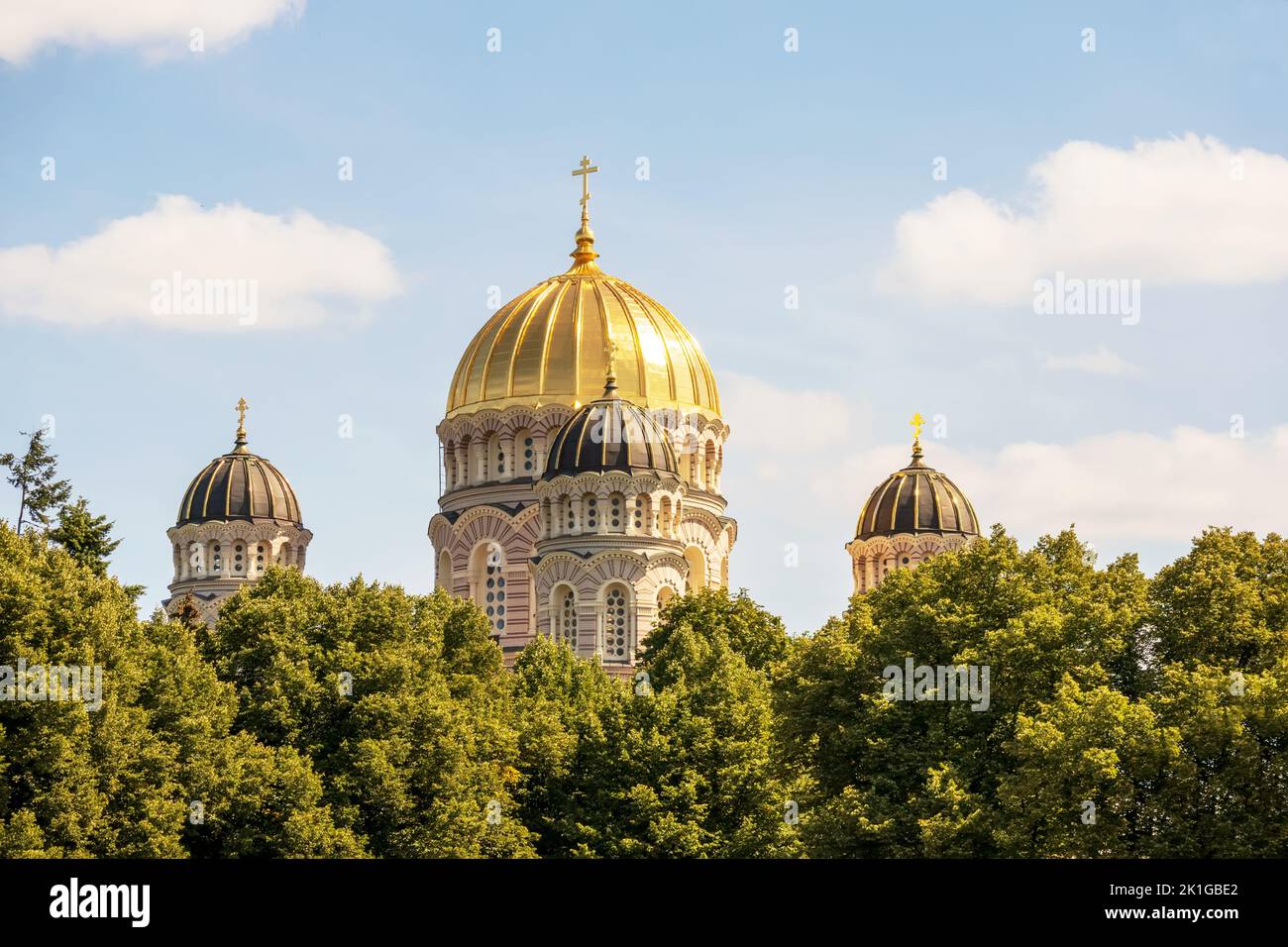 A church with a yellow wall and a pointed golden tip in the middle of the city center with multiple windows and trees next to it Stock Photo