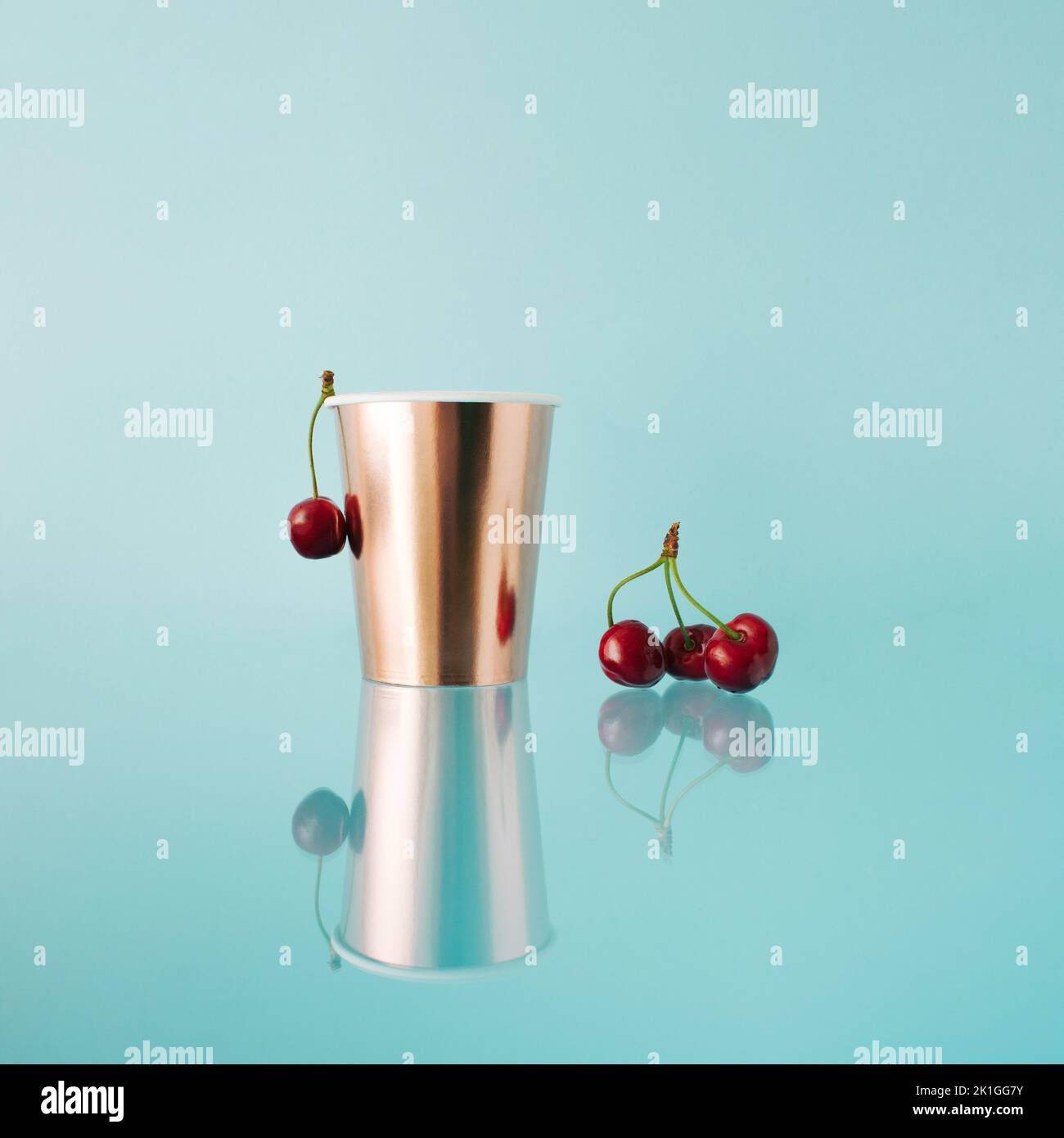 Golden paper cup with fresh cherries on blue reflecting background. Mirror reflection. Stock Photo