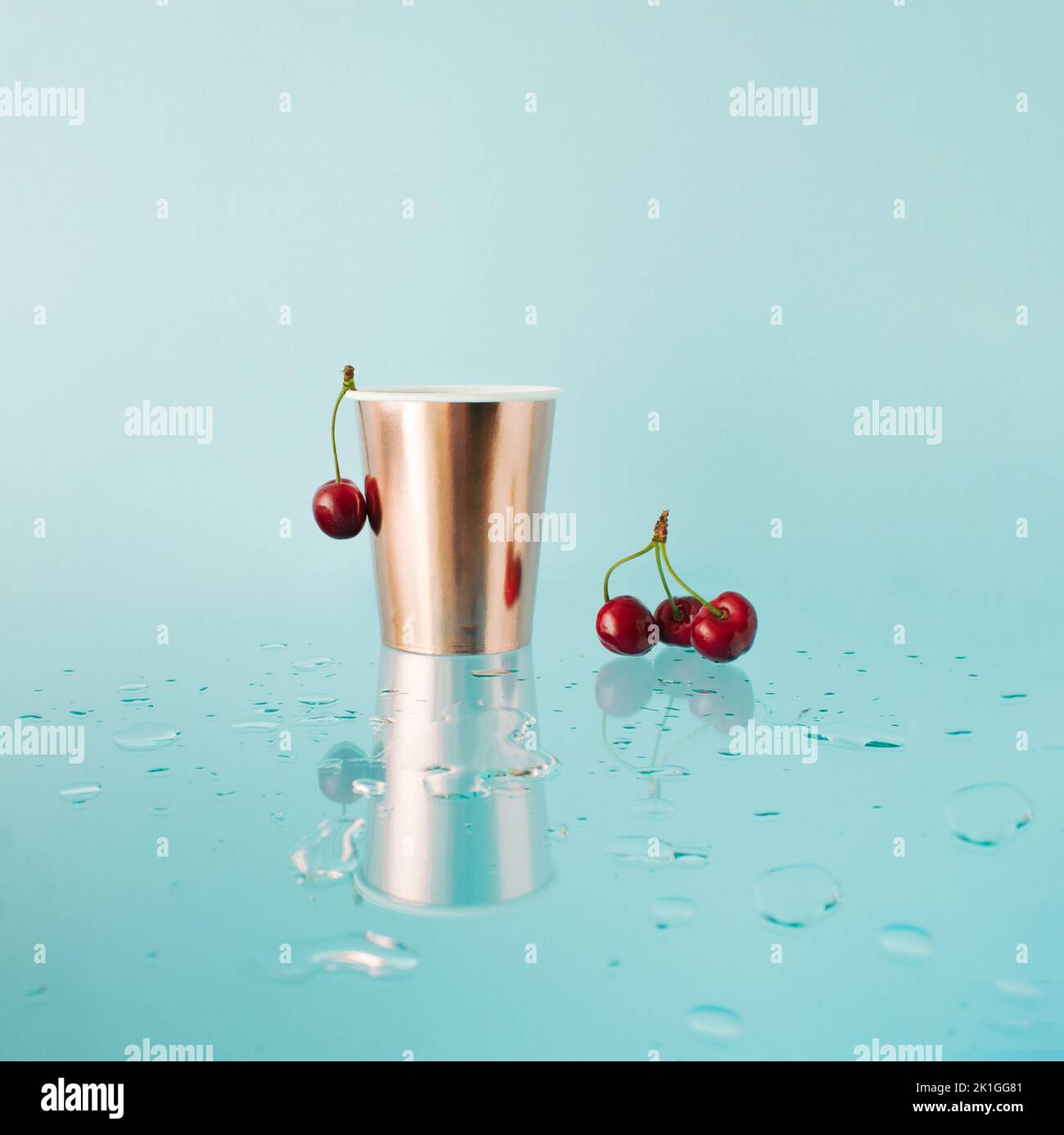 Golden paper cup with fresh cherries on blue reflecting background. Mirror reflection with drops of water. Stock Photo