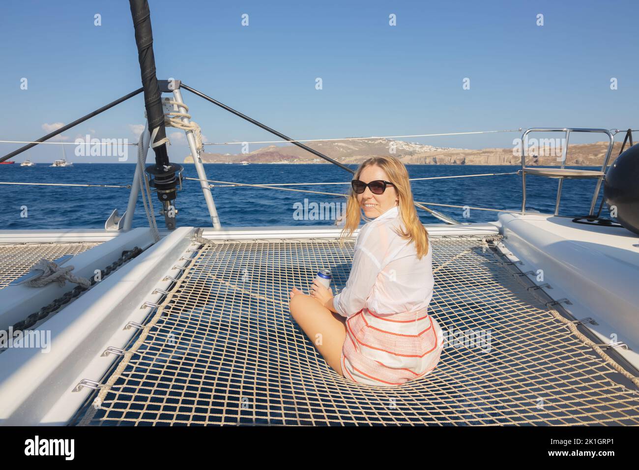 A young blonde female tourist enjoys a catamaran boat excursion on the Aegean Sea along the coast of the Greek Island Santorini, Greece. Stock Photo
