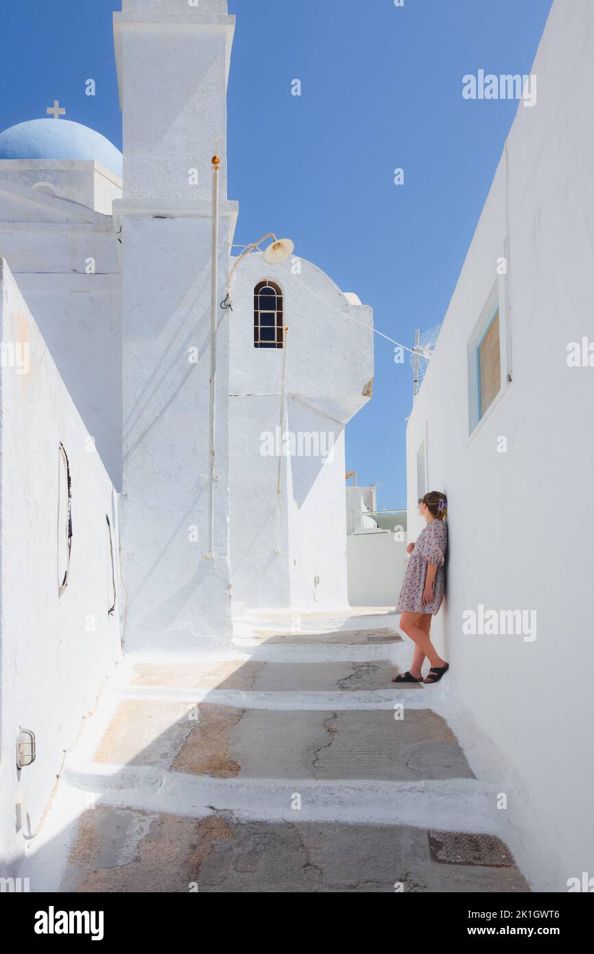 A young blonde female tourist leans up against a traditional white wash building while admiring a blue dome church in the quaint village of Pyrgos Kal Stock Photo