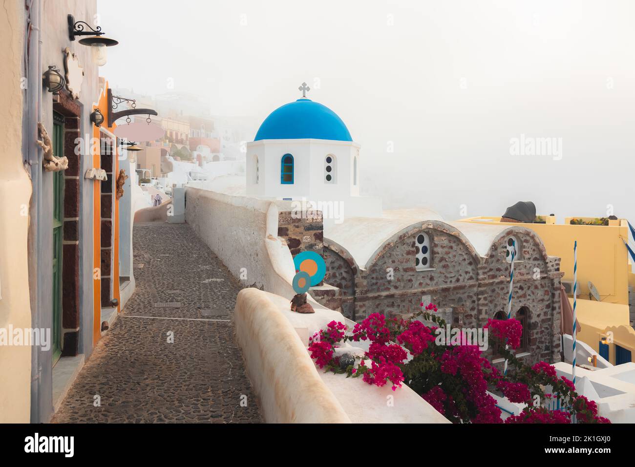A foggy, misty morning along a quiet, quaint cobblestone lane alongside the traditional blue dome Greek Orthodox church Panagia Agion Panton in the co Stock Photo