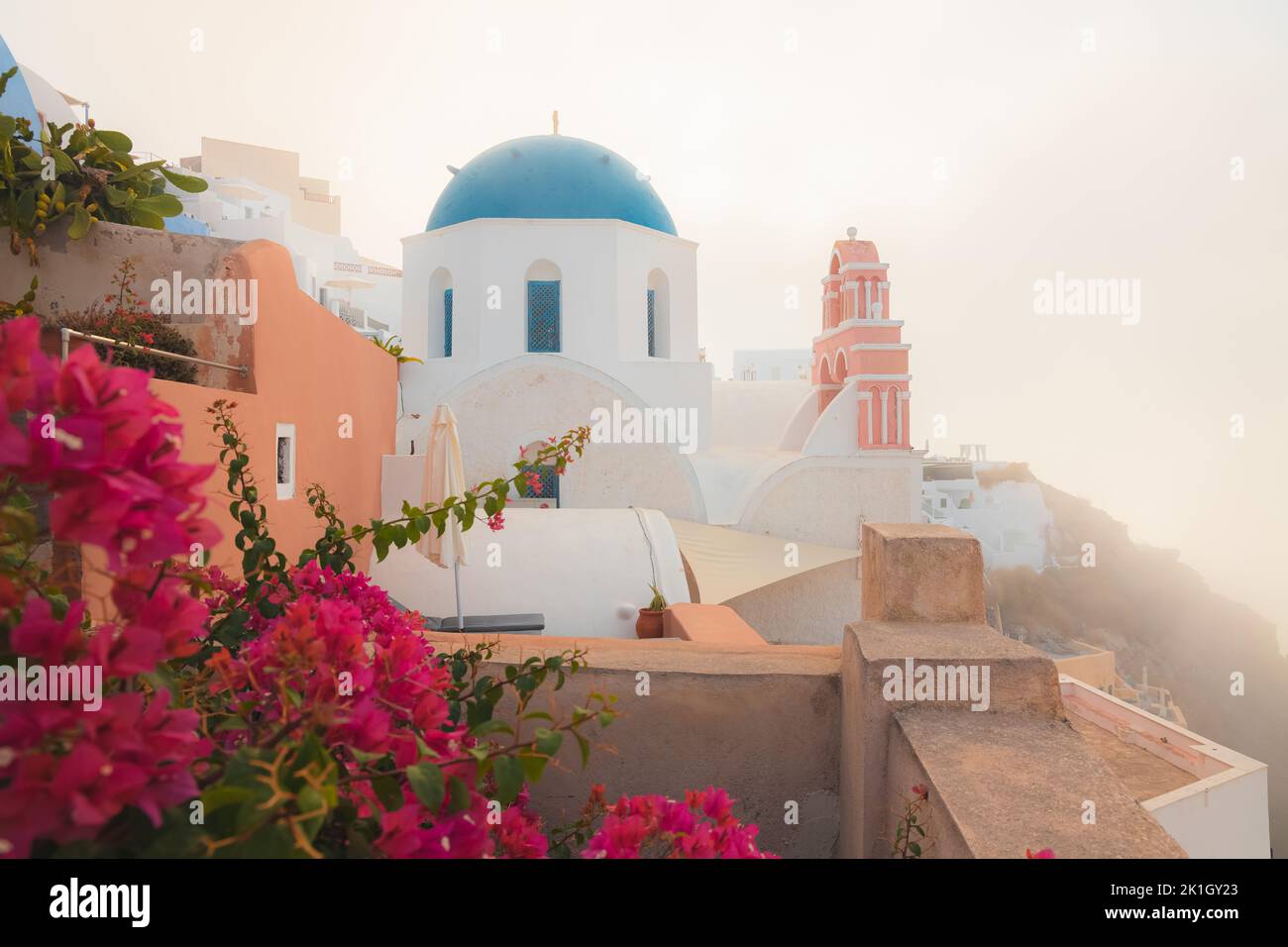 Bright red bougainvillea flowers on a foggy, misty morning during golden hour sunrise at the traditional blue dome Greek Orthodox Church of Agios Niko Stock Photo