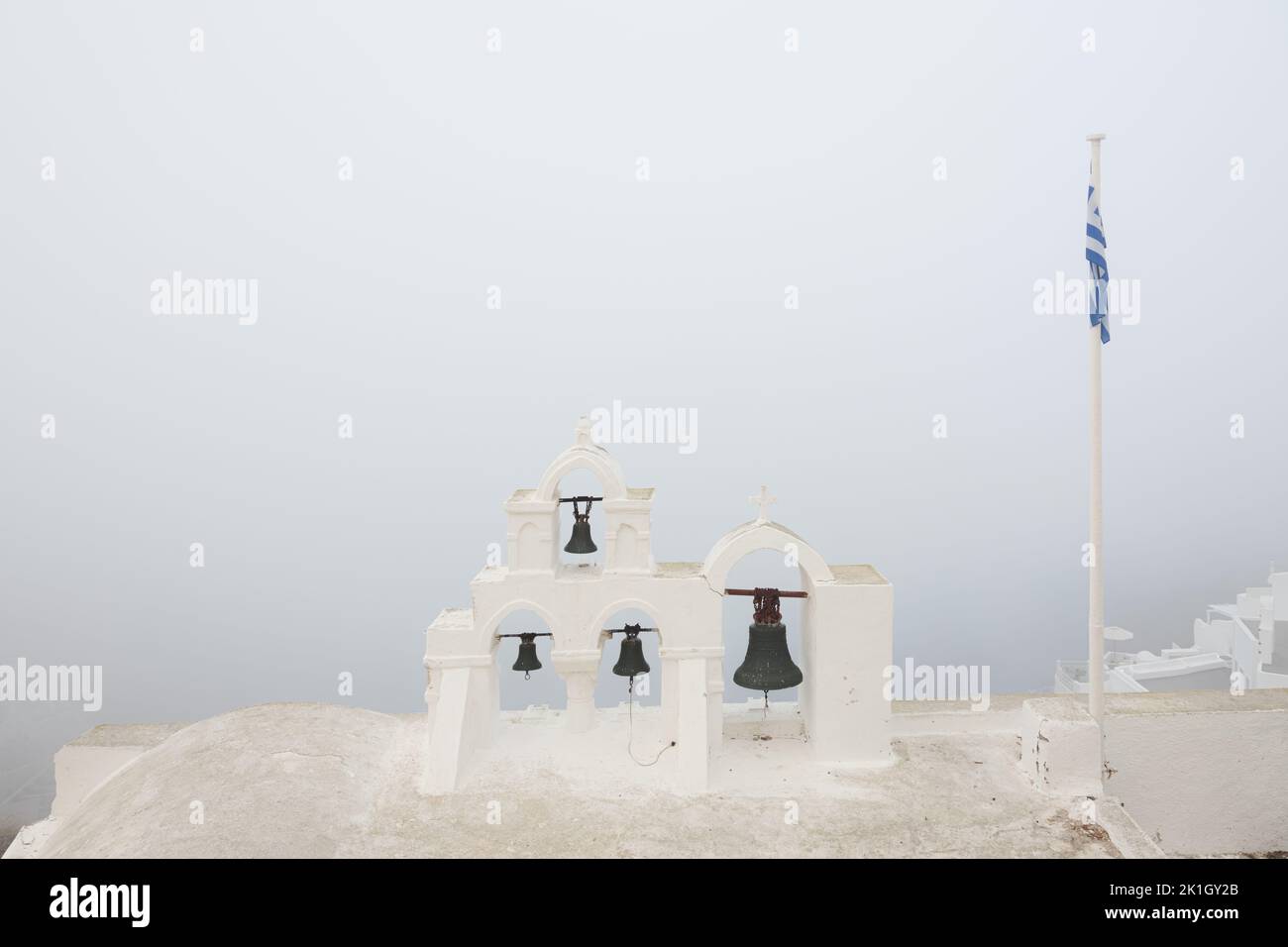 Minimalist scene of traditional Greek Orthodox church bells and Greek flag on a moody, atmospheric morning of fog and mist at the coastal village old Stock Photo