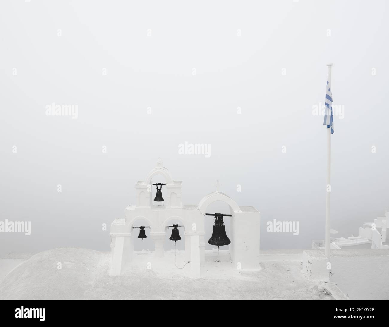 Minimalist scene of traditional Greek Orthodox church bells and Greek flag on a moody, atmospheric morning of fog and mist at the coastal village old Stock Photo