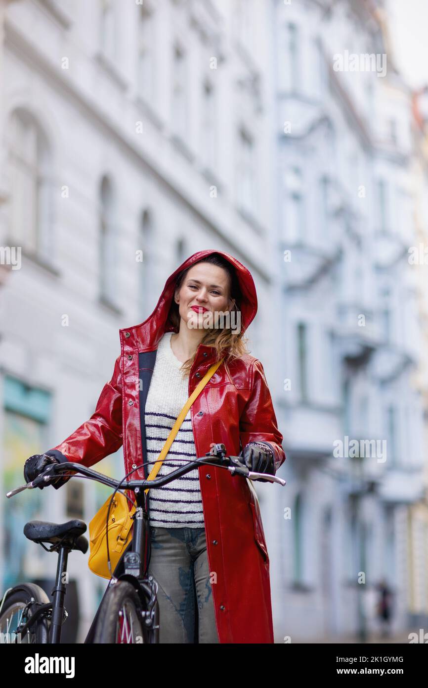 happy stylish female in red rain coat with bicycle in the rain outdoors on the city street. Stock Photo