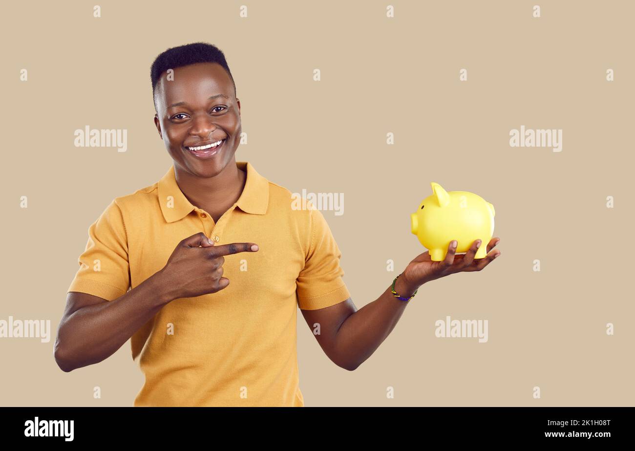 Young African American man points with finger and holds piggy bank for money on beige background Stock Photo
