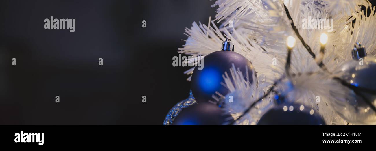 Wide view image of shiny blue holiday baubles hanging on white christmas tree with lights. With copy space. Stock Photo