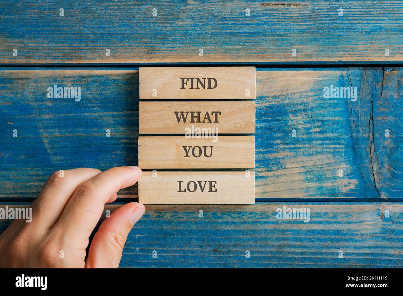 Male hand assembling a Find what you love sign written on four stacked wooden pegs placed on textured blue wooden boards. Stock Photo