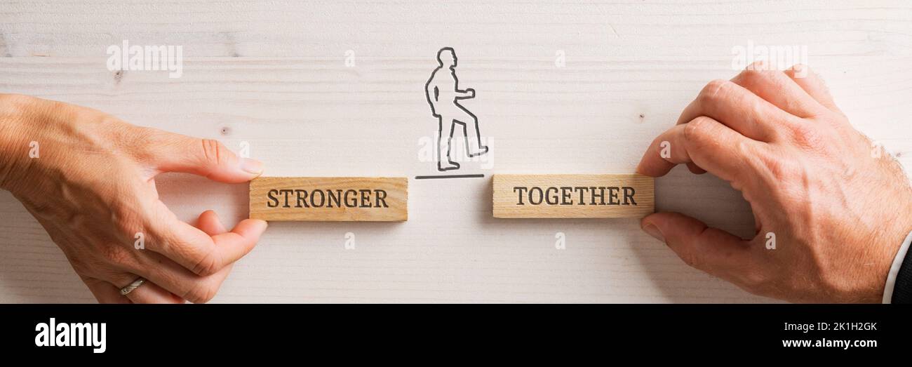Hands of a businessman and businesswoman holding two wooden pegs with a Stronger together sign for a silhouetted businessman to walk confidently betwe Stock Photo
